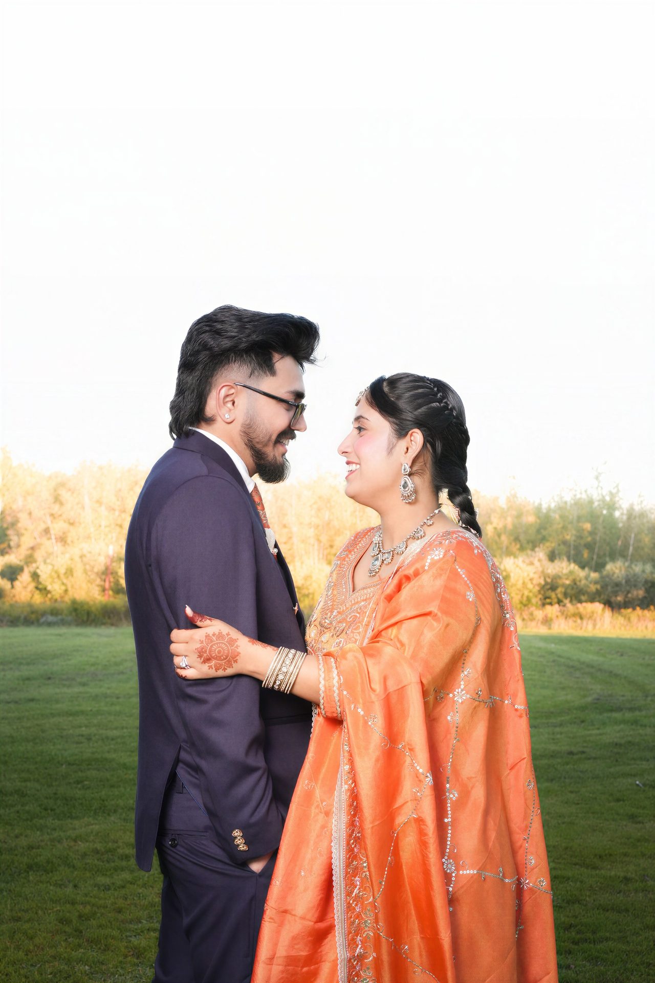 A couple in an embroidered orange outfit and a navy suit stand close and look at each other, a glowing field behind them.