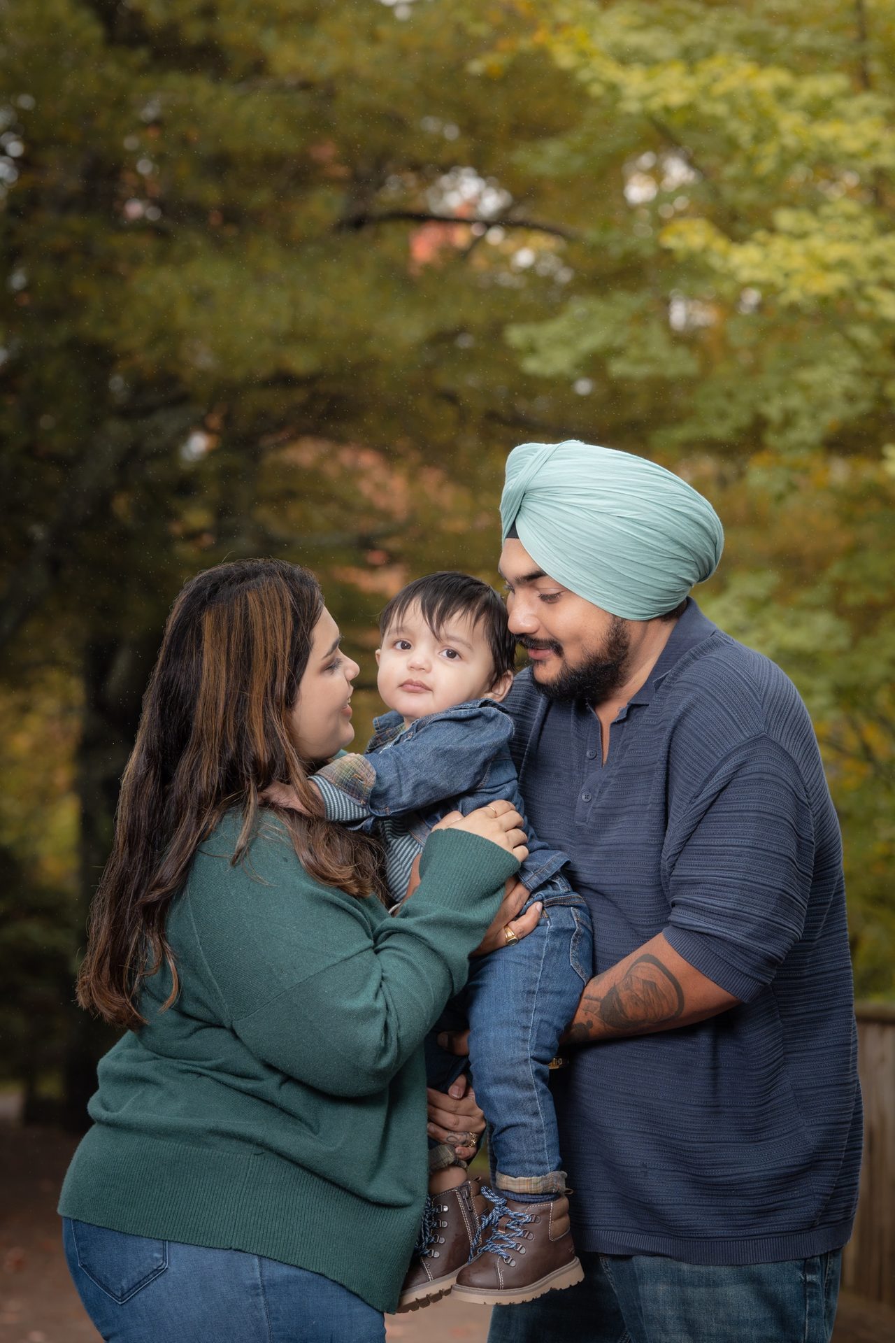 A woman in a green sweater and a man in a pale blue turban hold a toddler between them and smile, autumn leaves behind.