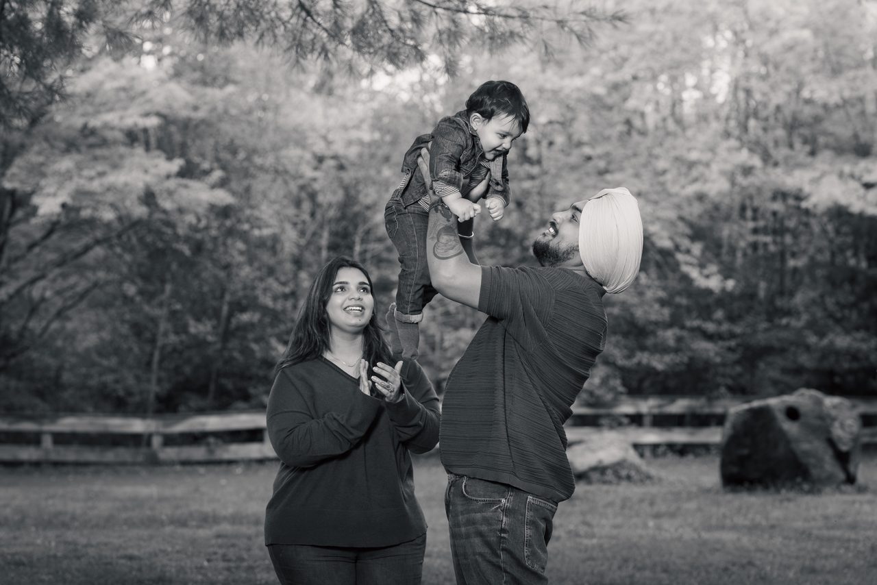 Black-and-white image of a man in a turban lifting a laughing toddler into the air while a woman looks up and claps.
