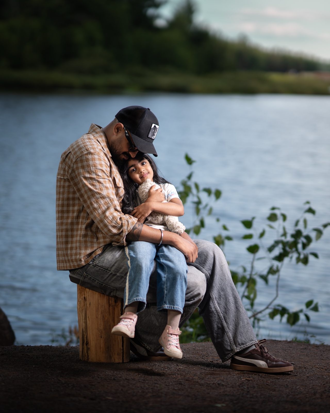 A man in a cap sits on a tree stump by a lake, holding a young girl who hugs a soft toy.