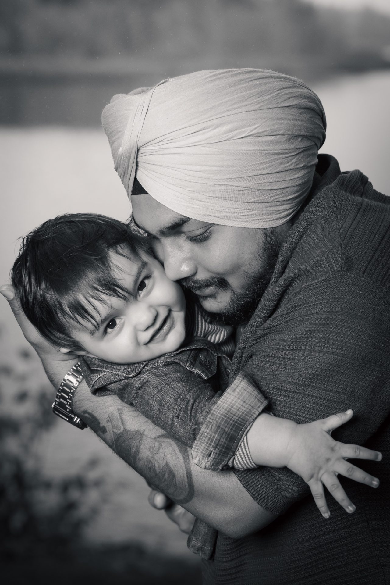 Black-and-white close portrait of a man in a turban hugging a smiling toddler beside a lake.