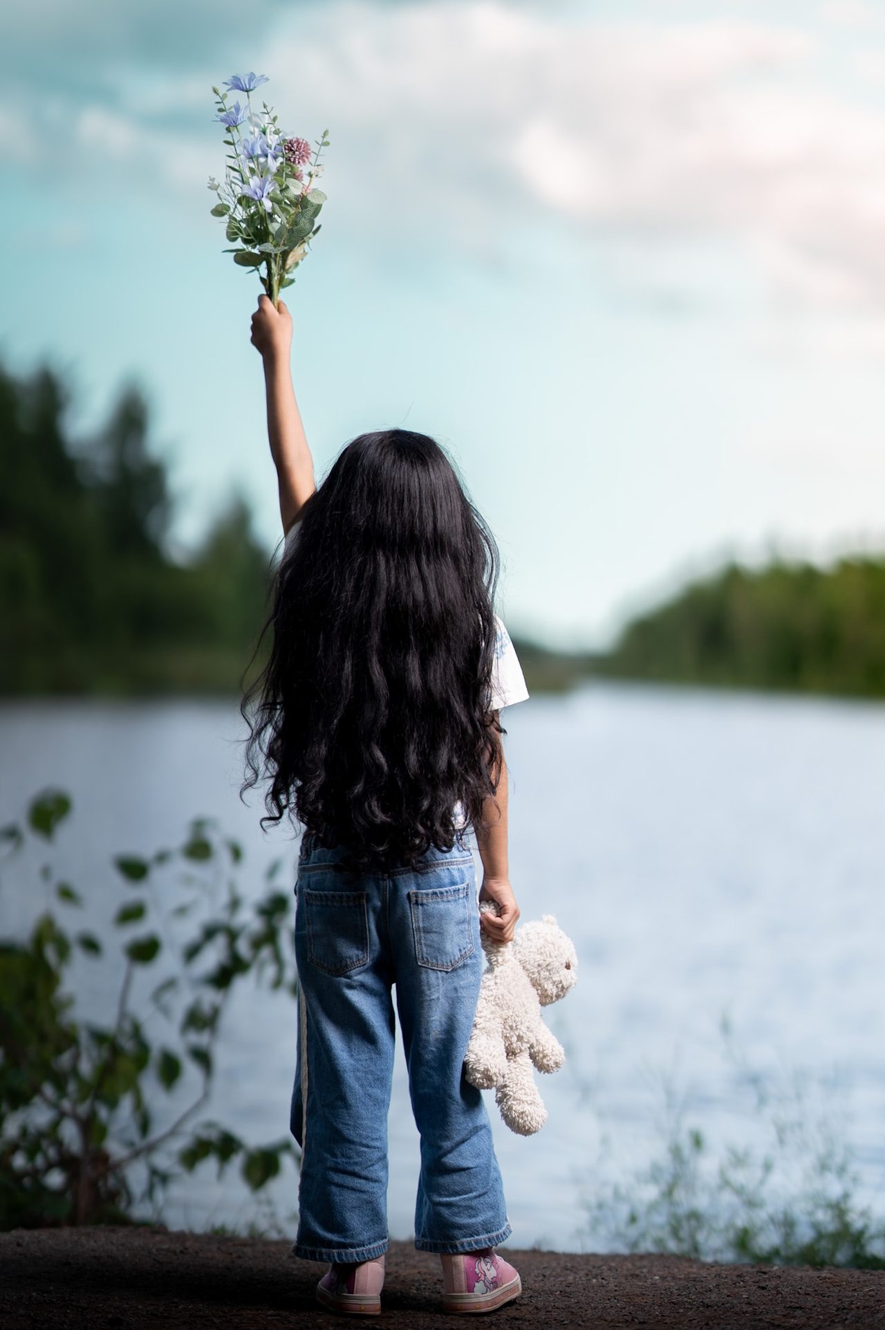 A young girl with long dark hair, seen from behind, reaches up with a bunch of flowers beside a lake, a soft toy in her other hand.