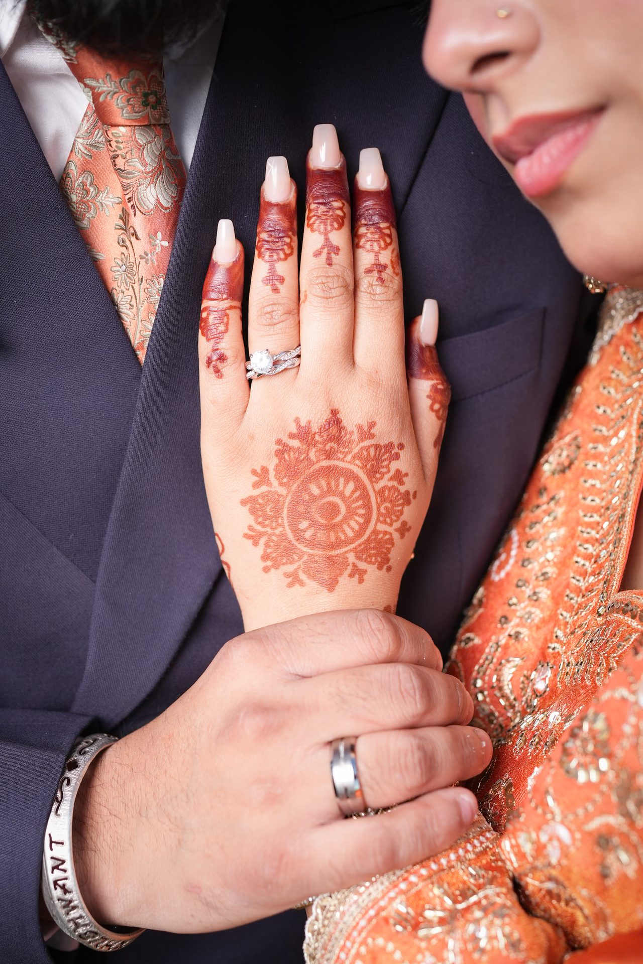 Close detail of a hand with a floral henna design and a diamond ring resting on a navy suit, held by another hand.