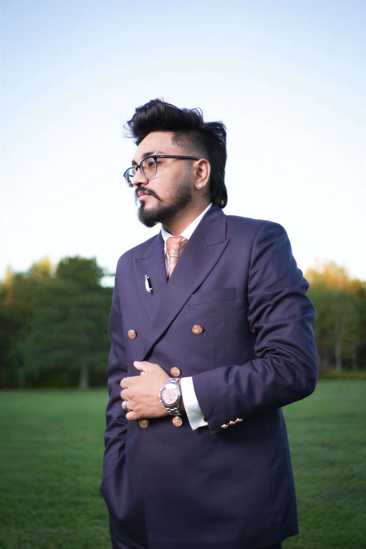 A man in a navy double-breasted suit with gold buttons looks off to the side on a lawn, trees and soft sky behind him.
