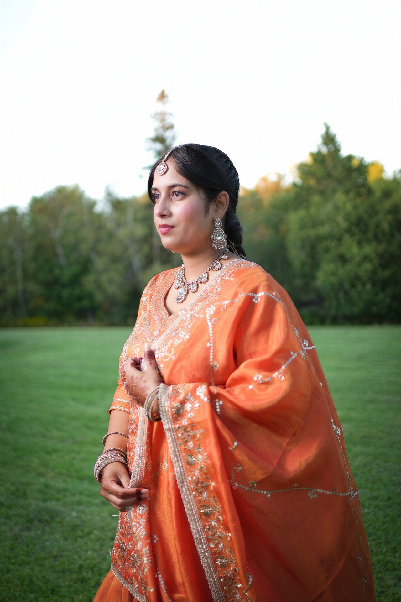 A woman in an embroidered orange outfit and traditional jewellery stands on a lawn and looks off to one side.